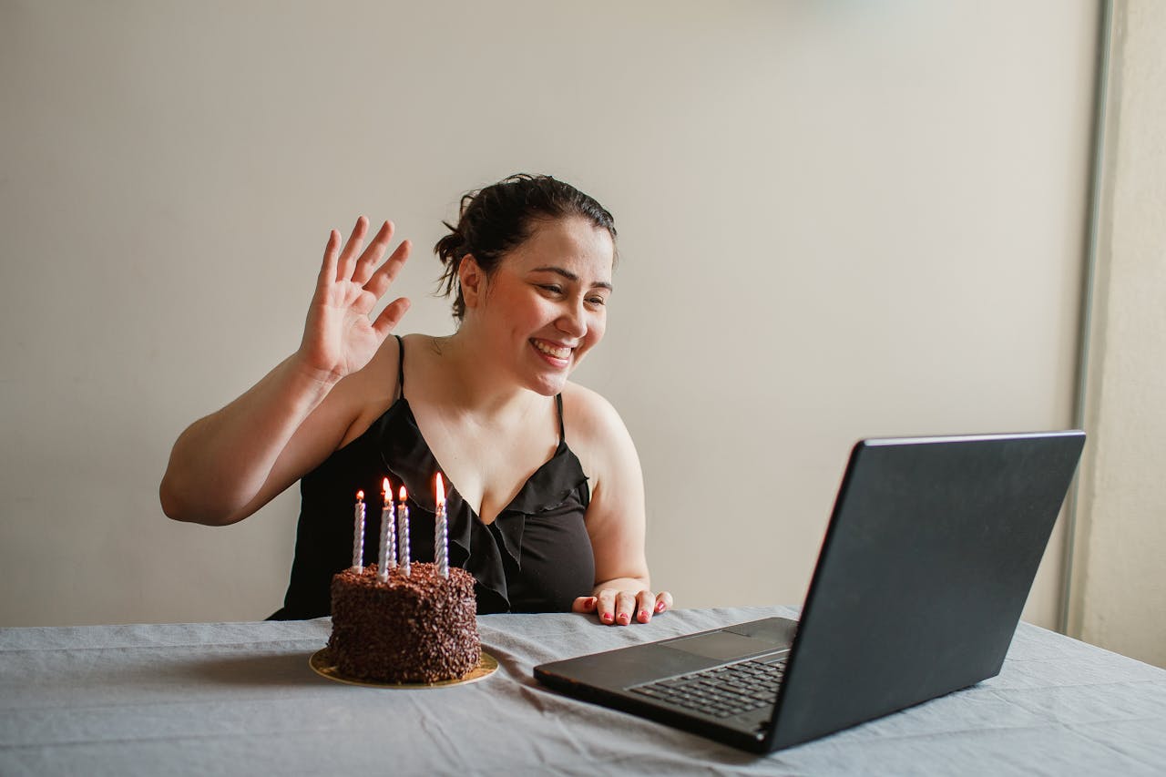A woman enjoys a virtual birthday with a cake, candles, and a laptop, celebrating online.